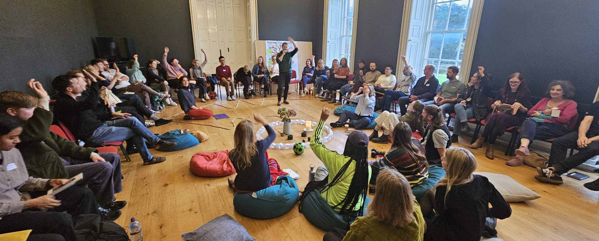 Philip standing inside a large circle of seated people in a panelled room, several hands raised.